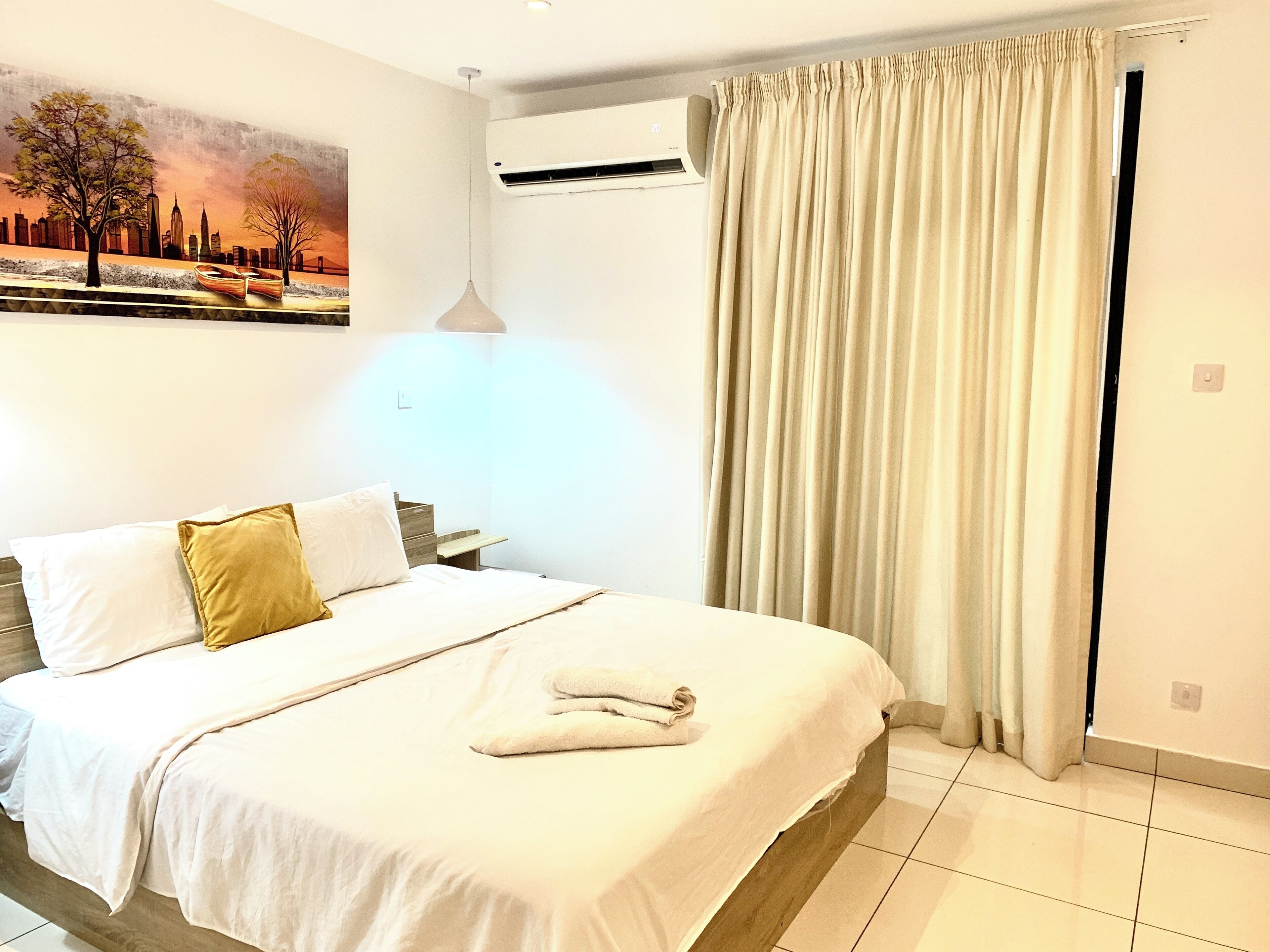 Une chambre d’hôtel avec un lit, un tableau représentant un paysage urbain, un appareil de climatisation et un rideau.