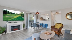 A modern living room with a fireplace, a flat-screen TV, and a dining area with blue chairs. - Pinehurst 2-bedroom, 2-bath Condo Available for Rent (Pinehurst)