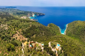 An aerial view of a coastal area with a house surrounded by dense greenery, a clear blue sea, and a small beach.