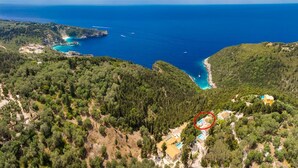 An aerial view of a coastal area with a small cluster of buildings surrounded by dense forest, overlooking a turquoise bay with boats.
