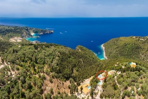 Eine Luftaufnahme einer Küstenlandschaft mit einer kleinen Gruppe von Gebäuden, umgeben von dichtem Wald, mit Blick auf eine türkisfarbene Bucht und darin liegende Boote.