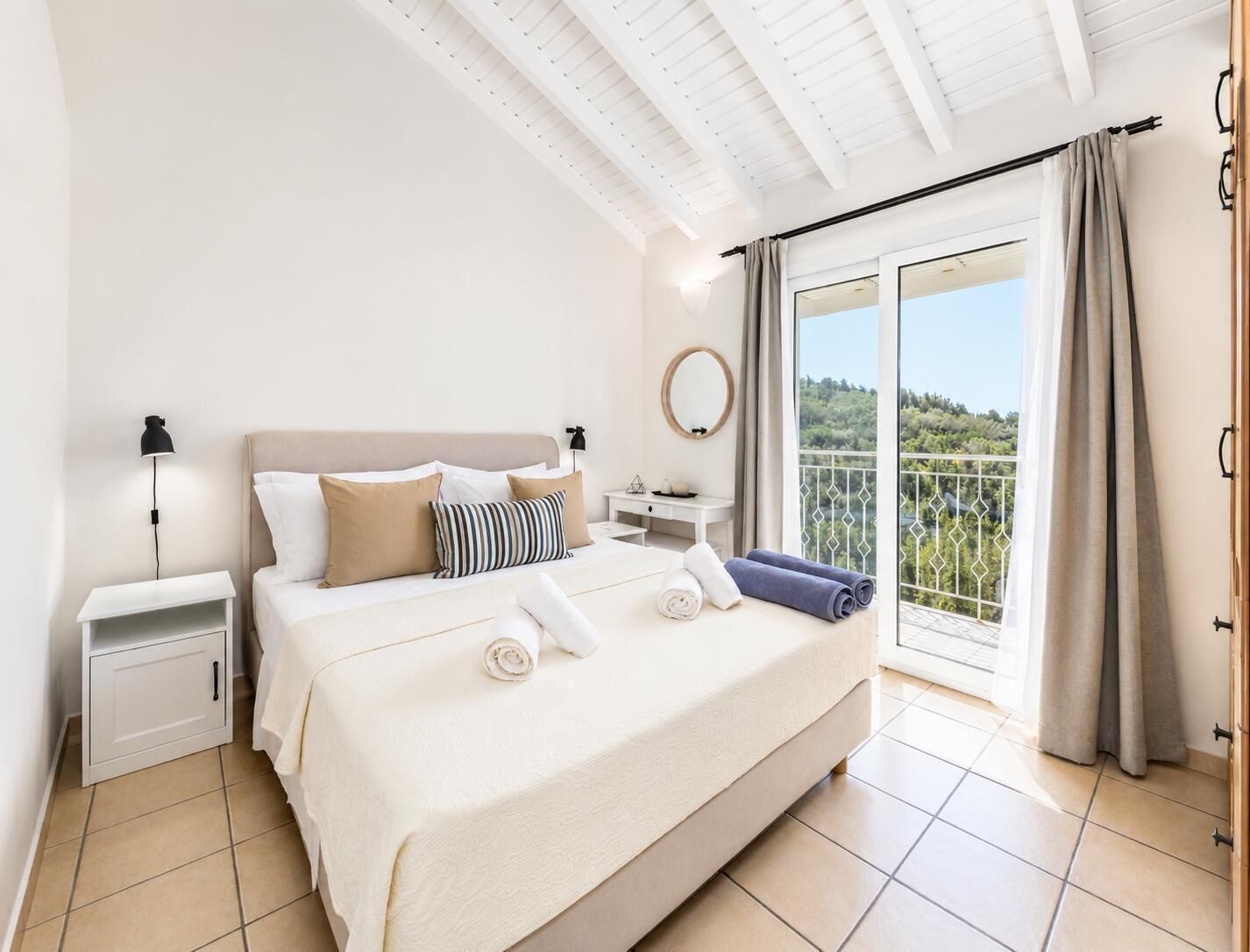 A bedroom with a bed, nightstands, a dresser, a round mirror, and a balcony with a view.