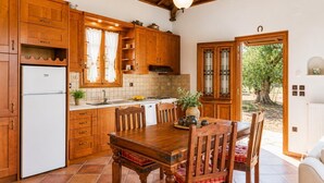 A kitchen with wooden cabinets, a white refrigerator, a dining table with chairs, and a view of trees outside.