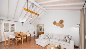 A spacious living area with a dining table, wooden chairs, a sofa, and a coffee table. There is a kitchen in the background with cabinets and a refrigerator. - Villa Gardenia - One Bedroom Villa, Sleeps 2 (Paxi)