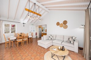 Zona d'estar àmplia amb taula de dinar, cadires de fusta, sofà i taula de centre. Es pot veure una cuina al fons amb armaris i nevera.