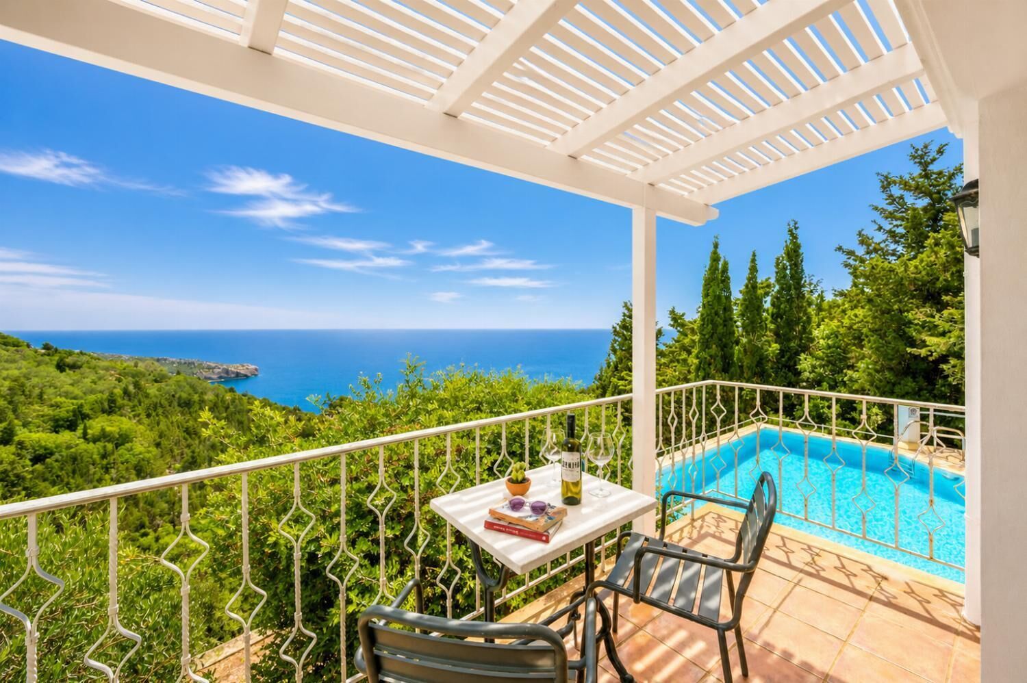 Un balcón con vista al mar, una piscina y una mesa con una botella y vasos.