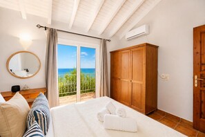 A bedroom with a bed, a wooden wardrobe, a mirror, a window with a view of the sea, and an air conditioning unit.