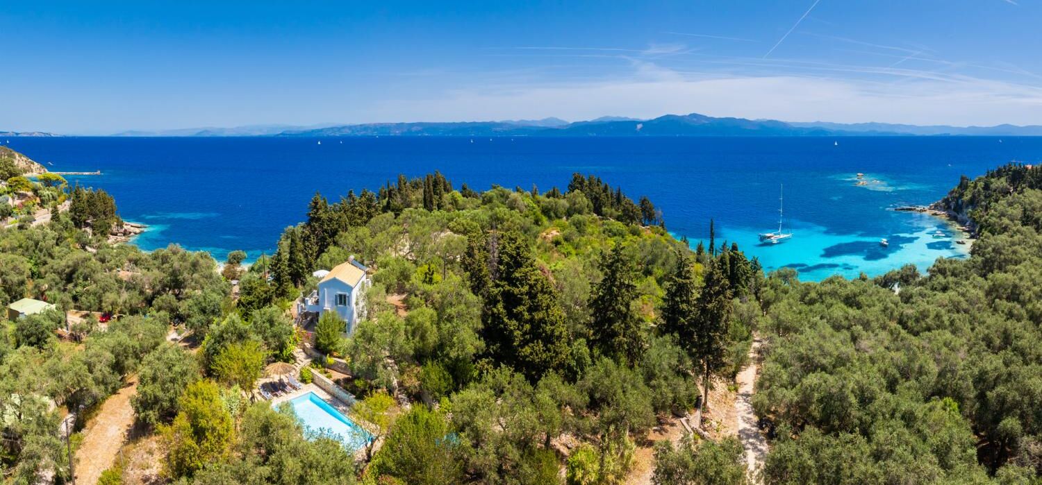 A coastal property with a swimming pool, surrounded by dense forest and overlooking a clear blue sea.