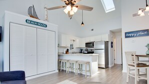 Una cocina moderna con armarios blancos, una isla central con taburetes, un ventilador de techo y un letrero que dice 'BEACH HOUSE'.