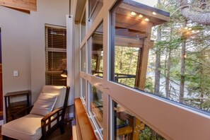 A cozy cabin interior with a wooden bench, a cushioned chair, and large windows offering a view of a forest and lake. - The Cove I Water Views I Wild Pacific Trails (Ucluelet)