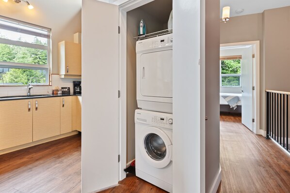 A laundry room with a washing machine and dryer, wooden cabinets, and a window with a view of greenery. - The Cove I Water Views I Wild Pacific Trails (Ucluelet)
