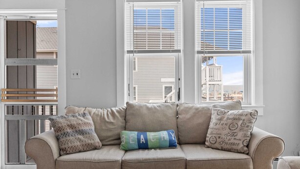 A beige sofa with three cushions, one with 'BEACHY' text, in a room with white walls and a view of a building through a window with blinds. - OCHG 6 10th Steet Top (Ocean City)