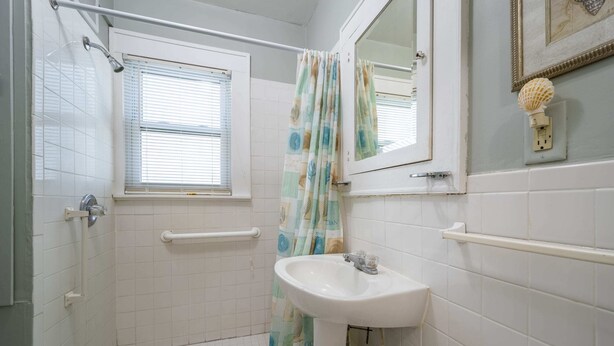 Un baño con ducha, lavabo, ventana con persianas, espejo y toallero.
