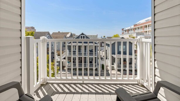 A balcony with a white railing, wooden deck, and a view of a residential area with houses and a building with a red roof.