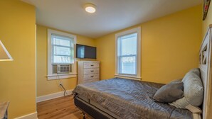 Une chambre à coucher comprenant un lit, une commode, un téléviseur et deux fenêtres équipées de stores.