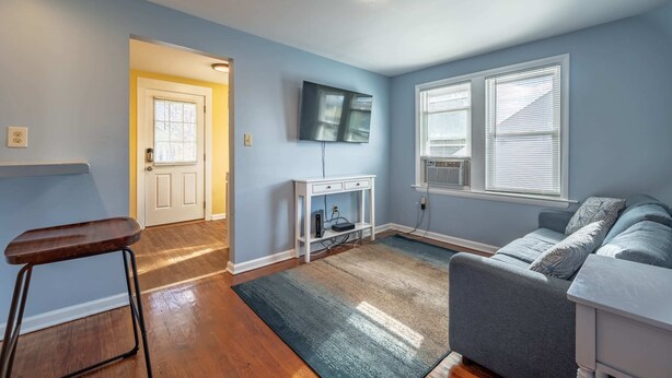 A living room with a grey sofa, a wooden floor, a white TV stand, and a door leading to another room.