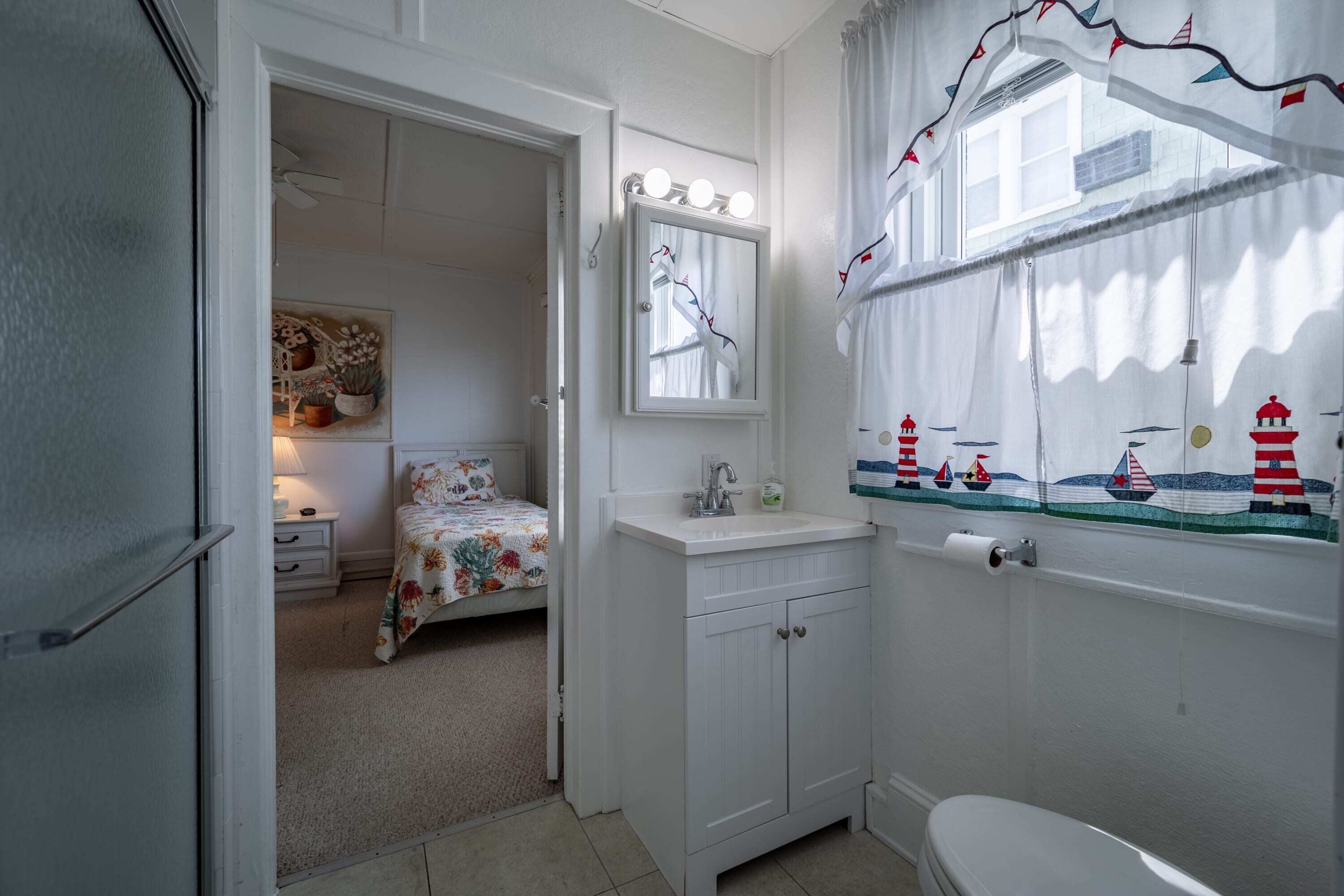 Een badkamer met een wit wastafel, spiegel en toilet. In de aangrenzende kamer staat een bed met bloemenbeddels.