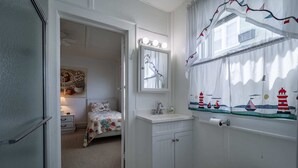 Een badkamer met een wit wastafel, spiegel en toilet. In de aangrenzende kamer staat een bed met bloemenbeddels.