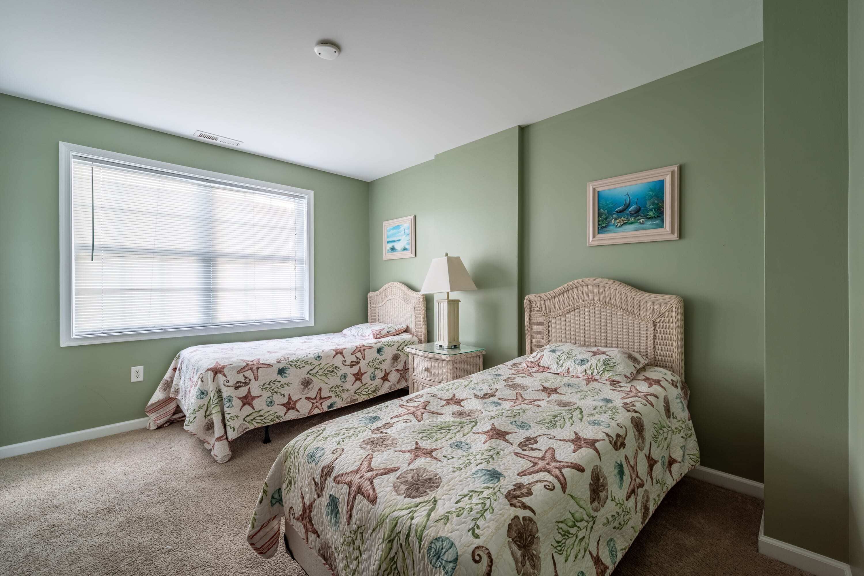 A bedroom with two beds, a window with blinds, a nightstand with a lamp, and two framed pictures on the wall.