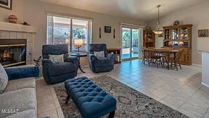 A spacious living room with a fireplace, two armchairs, a blue ottoman, and a dining area with a wooden table and chairs.