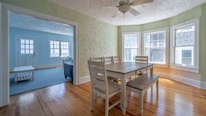 A dining area with a wooden table and chairs, a ceiling fan, and large windows with white frames. - OCHG Seafoam 1005 Sum (Ocean City)