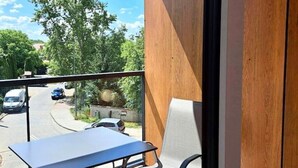 Ein Balkon mit Tisch und Stuhl, der auf eine Straße mit parkenden Autos und Bäumen blickt.