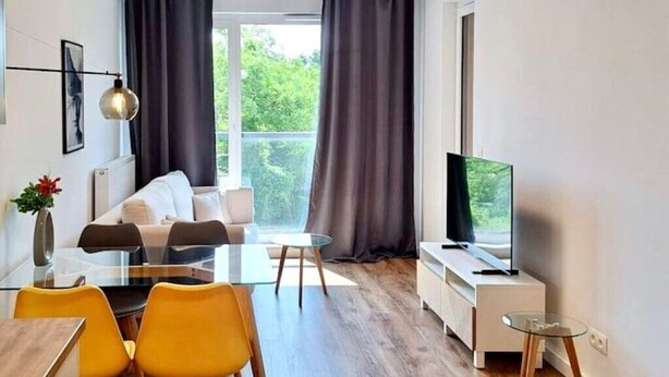 Ein modernes Wohnzimmer mit einem Glastisch, gelben Stühlen, einer Couch, einem Fernseher und Blick auf Grünpflanzen durch große Fenster.