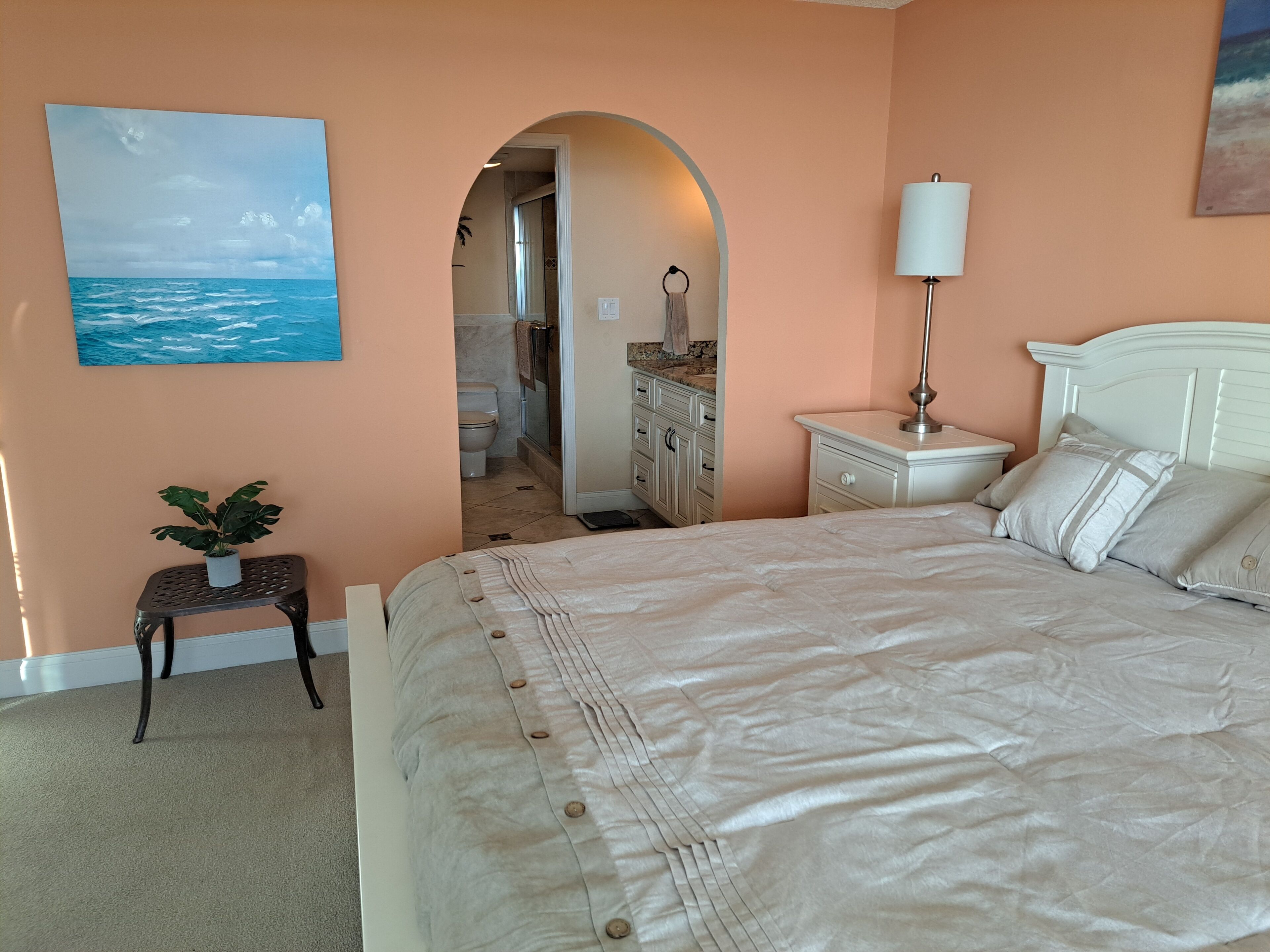A bedroom with a large bed, a nightstand, a lamp, a small table with a plant, and a painting of the ocean.
