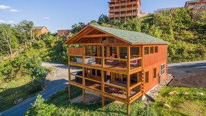 A multi-story wooden cabin with balconies, situated on a hillside with a view of a larger building in the background. - The Sweetest Cabin In The Smokies (PIGEON FORGE)