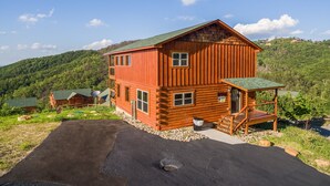 A log cabin with a green roof, a porch, and a view of the surrounding forest. - The Sweetest Cabin In The Smokies (PIGEON FORGE)