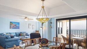 Ein Wohnzimmer mit einem blauen Sofa, einem Esstisch mit Stühlen, einem Deckenventilator und Blick auf den Ozean durch eine Schiebetür.