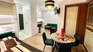 Ein modernes Wohnzimmer mit einer Couch, einem Esstisch mit Stühlen, einem an der Wand montierten Fernseher und einer Küche im Hintergrund.