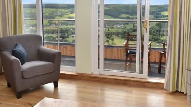 A room with a grey armchair and a view of a green landscape through large sliding glass doors. - Glen View Lodge (Inverness)