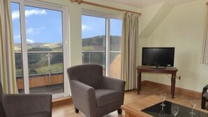 A living room with a sofa, a coffee table with wine glasses, a television on a stand, and large windows with a view of the countryside. - Glen View Lodge (Inverness)