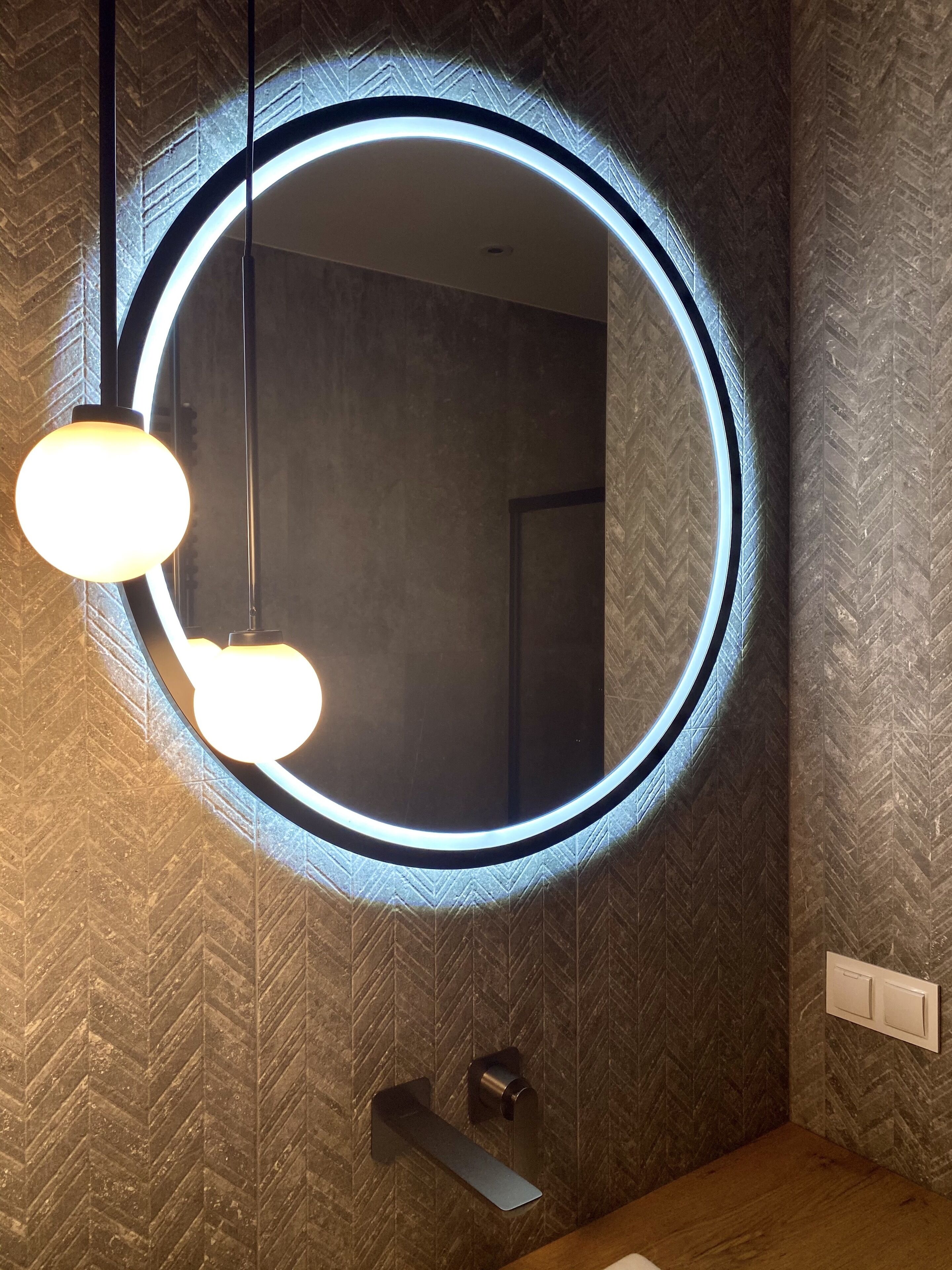Une salle de bain avec un miroir rond, deux lampes suspendues et un mur à texture.