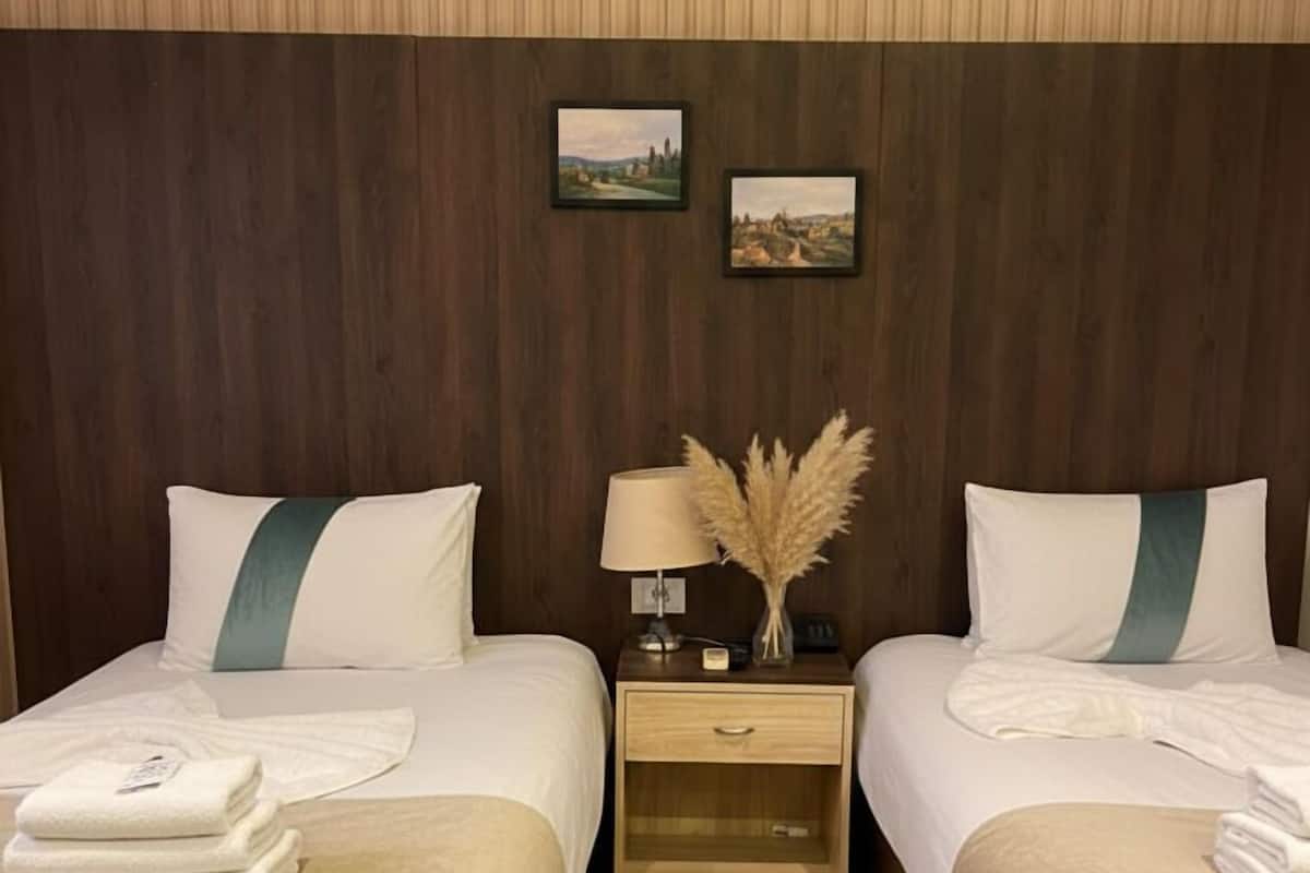 Habitación de hotel con dos camas, cada una con cubrecama y almohadas blancas, una mesita de noche con lámpara y objetos decorativos, y paneles de madera en las paredes.