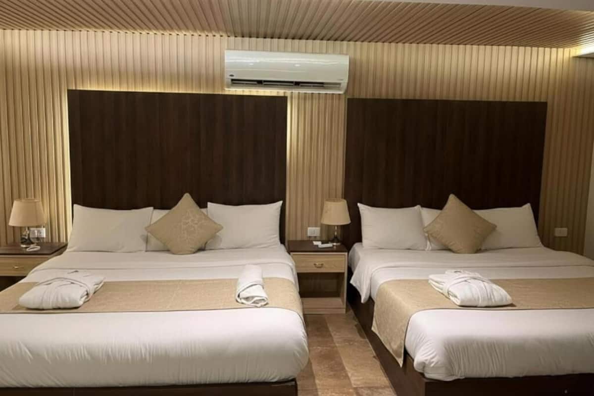 Habitación de hotel con dos camas, paneles de madera, unidad de aire acondicionado y una mesita de noche con lámpara.