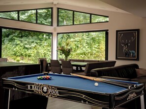 A pool table with a 'BALL POOL' label, a sofa, and a framed deer picture in a room with large windows.