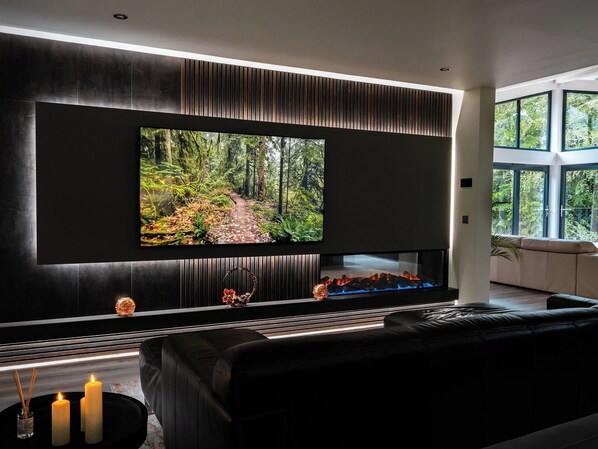 A modern living room with a large flat-screen TV mounted on a dark wall, a black leather sofa, and a fireplace with a glass front.