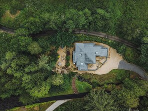 An aerial view of a house surrounded by dense greenery, with a clear driveway leading to the entrance.