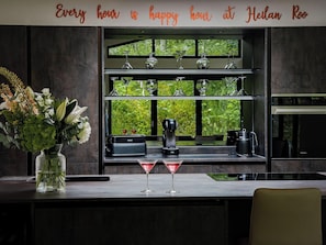 A bar area with a glass vase of flowers, a martini glass, a coffee machine, and a shelf with glasses.