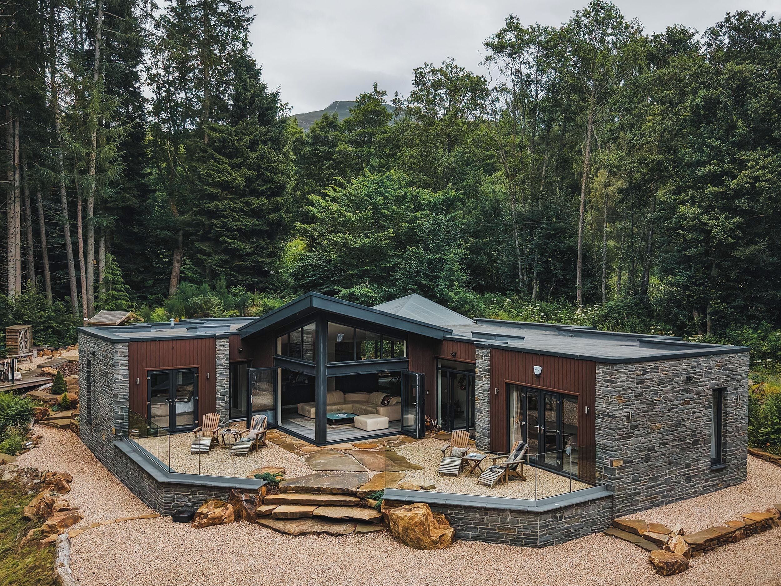 Une maison moderne avec une façade en pierre, de grandes fenêtres en verre et un patio doté d’un espace salon extérieur.