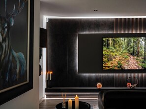 A modern living room with a large flat-screen TV mounted on a dark wall, a framed painting of a deer, a small table with candles, and a decorative item resembling a tree branch. - Heilan Roo (Pitlochry)