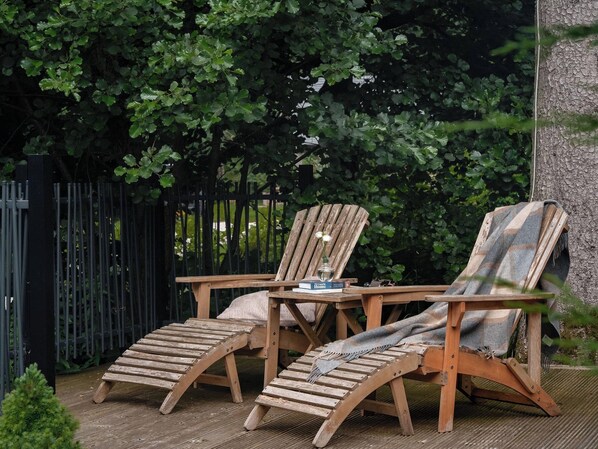 Two Adirondack chairs with a small table and a book on it, set on a wooden deck with a black metal fence in the background. - Heilan Roo (Pitlochry)