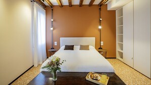 A hotel room with a large bed, wooden beams on the ceiling, a bedside table with a vase of flowers, and a tray with food and drinks. - Palazzetto San Stin (Venice)