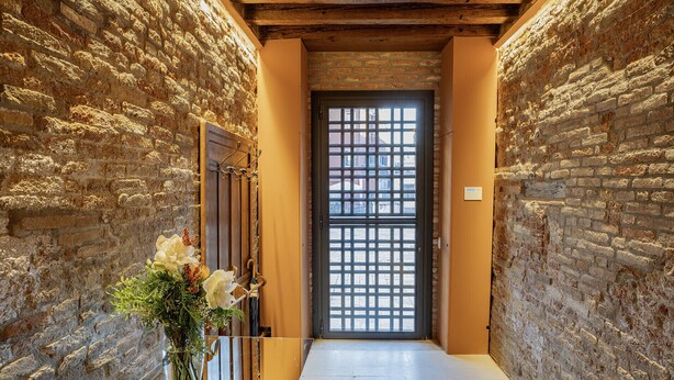 A narrow hallway with stone walls and a wooden door with a grid pattern. - Palazzetto San Stin (Venice)