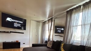 Ein modernes Wohnzimmer mit einem großen Flachbildfernseher an der Wand, einem dunkelgrauen Sofa mit gemusterten Kissen und einem grünen Hocker.
