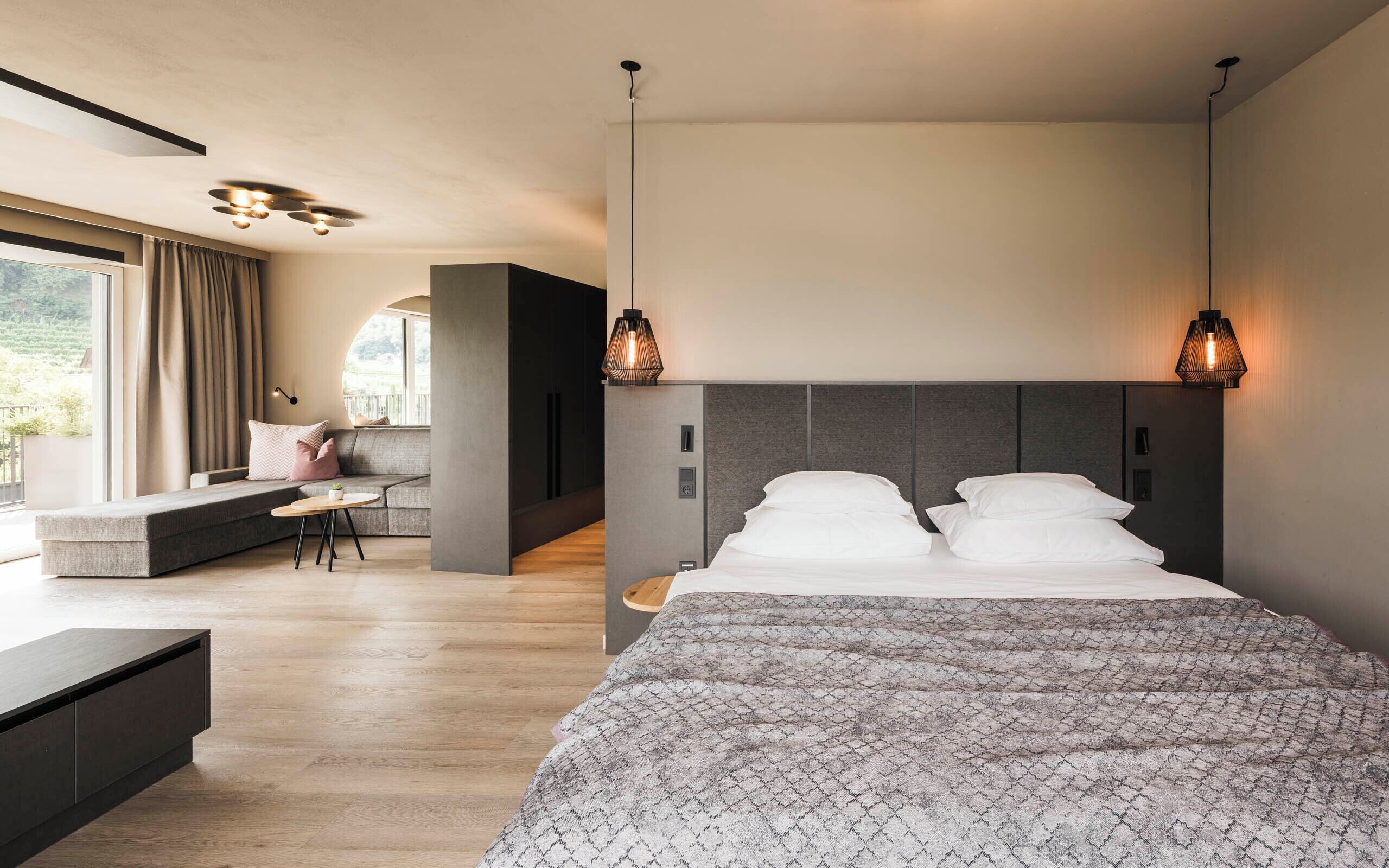 A modern bedroom with a large bed, a seating area with a sofa and a small table, and a view of the outdoors through a large window.