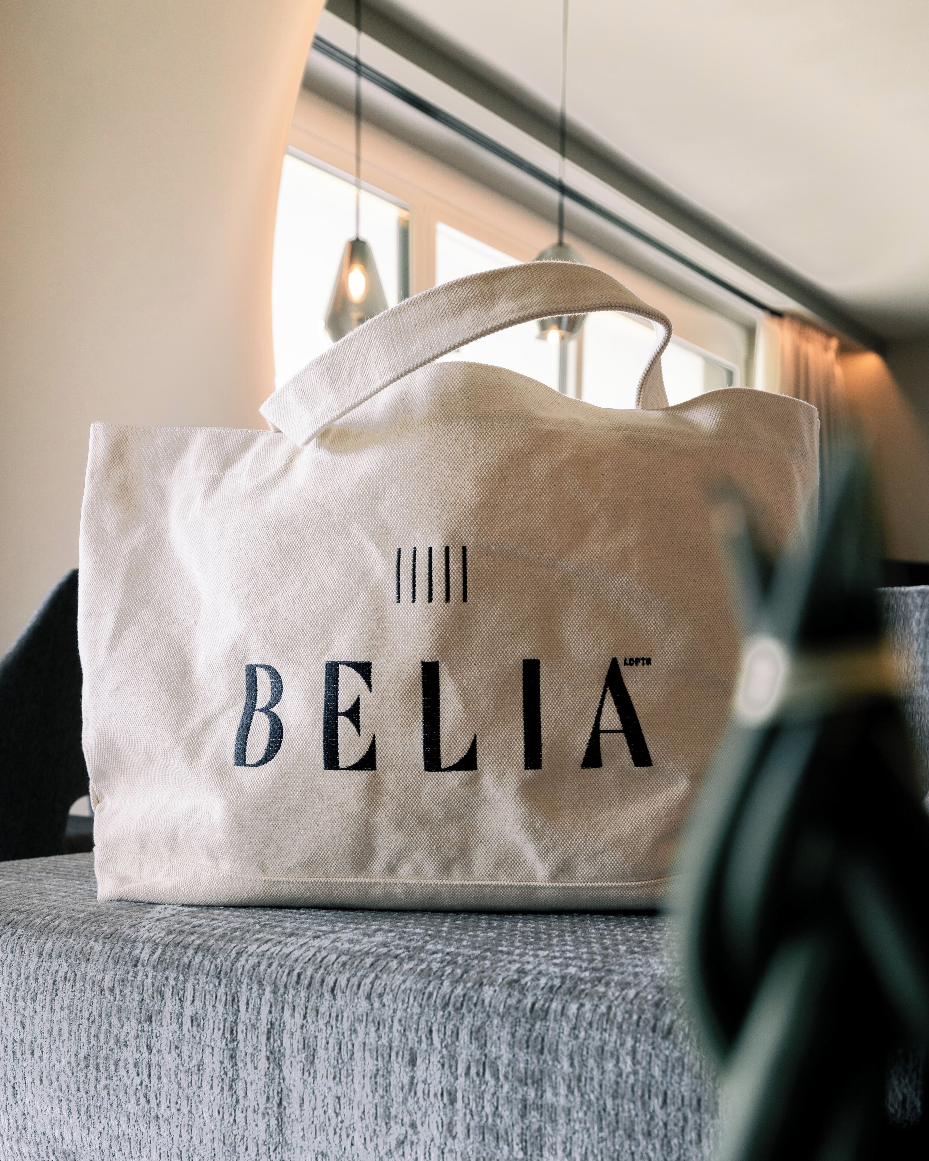 Uma bolsa branca de tela com a palavra 'BELIA' bordada, colocada em um sofá cinza.