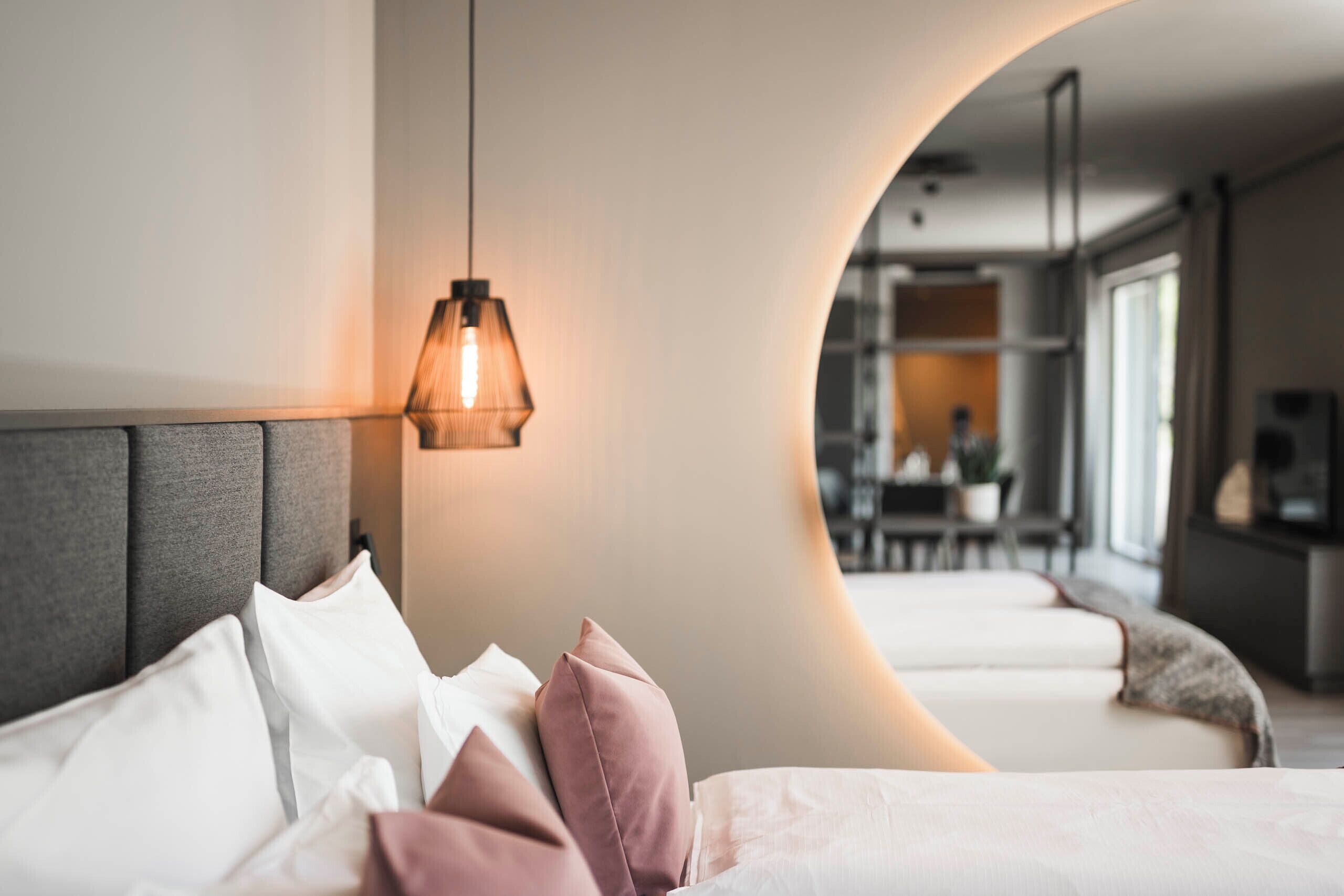A modern hotel room with a curved wall, a bed with white and pink pillows, a hanging light fixture, and a view into the dining area.